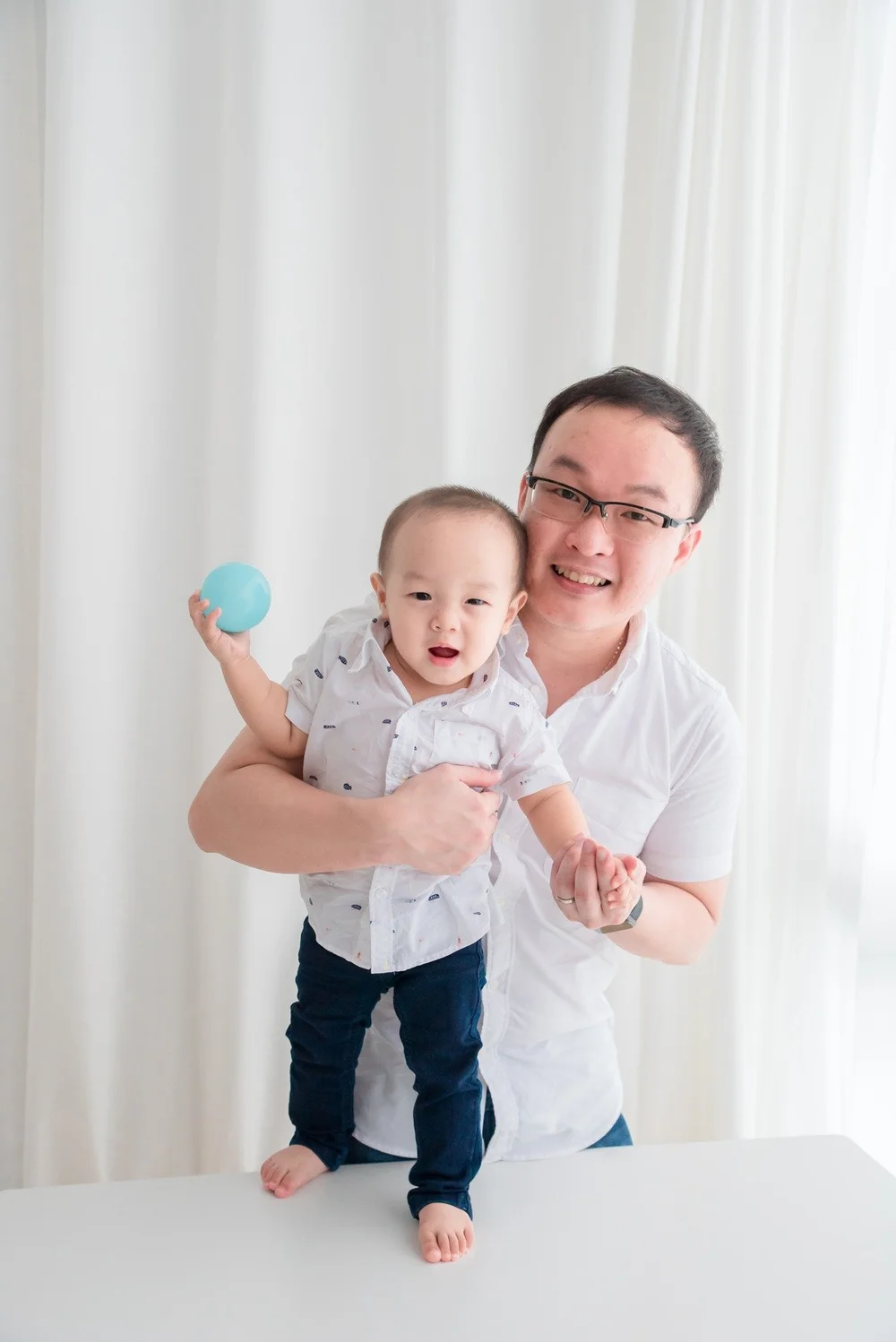 Smiling father supporting his baby as the baby holds a blue ball, photographed by Mamamiyo Photography in Singapore.
