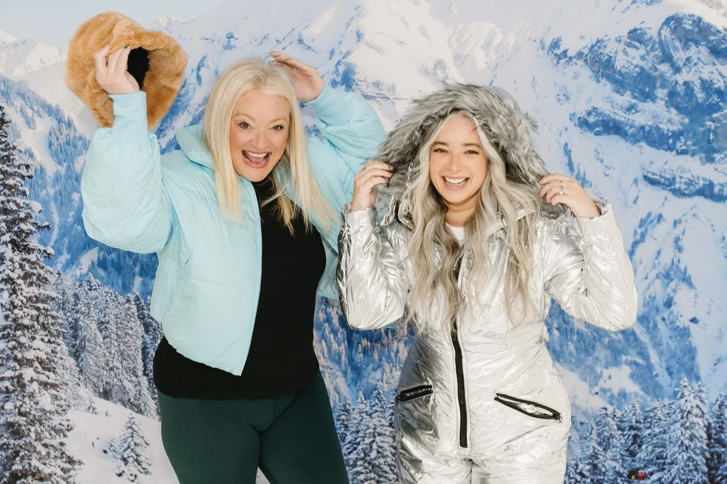 Two guests in ski jackets laughing at a winter-themed holiday party photo booth backdrop