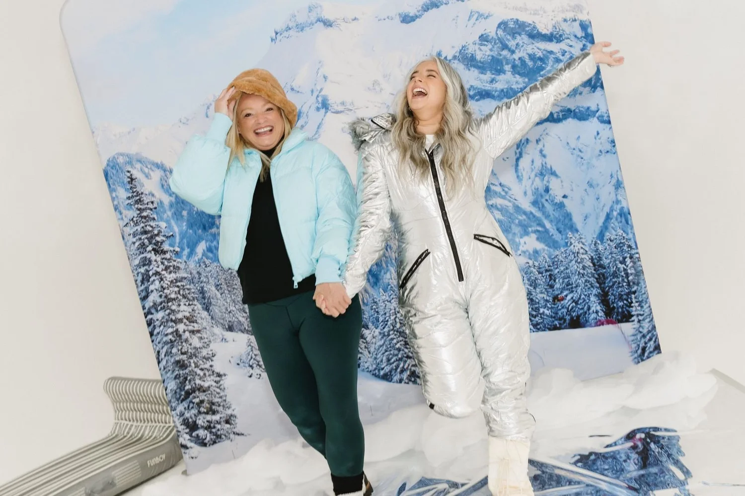 Two friends in puffer jackets laughing in front of a snowy mountain winter wonderland photo booth backdrop