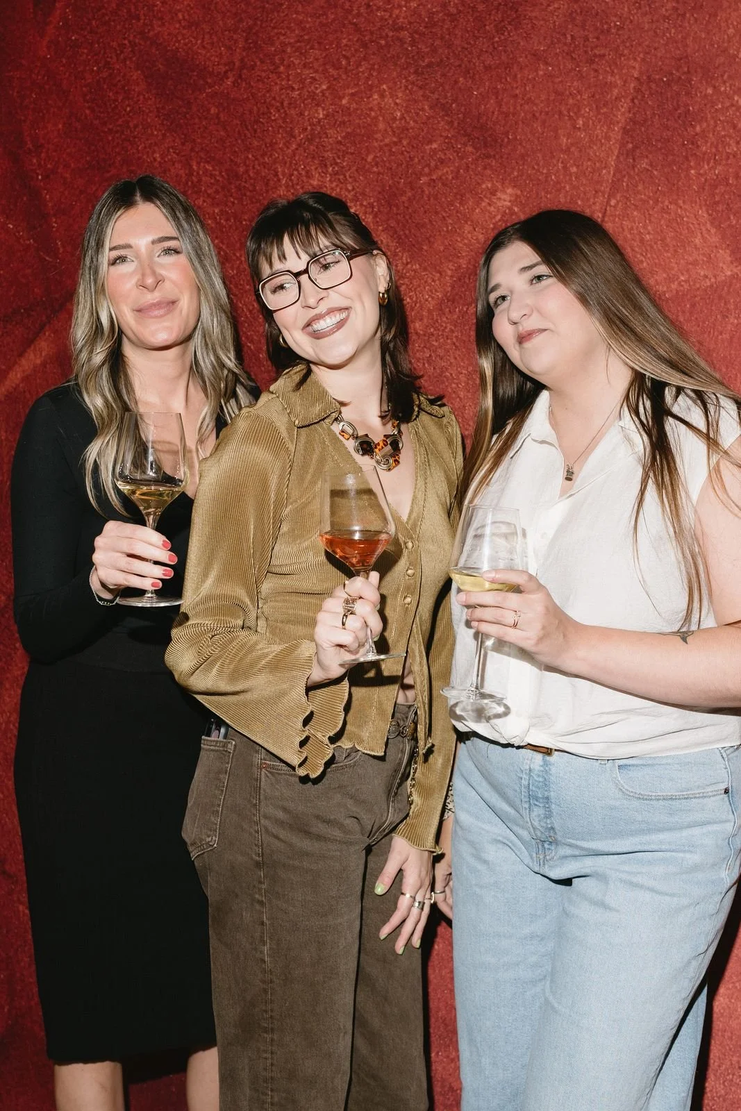 Three coworkers holding glasses of wine and laughing at a photo booth with a terracotta backdrop
