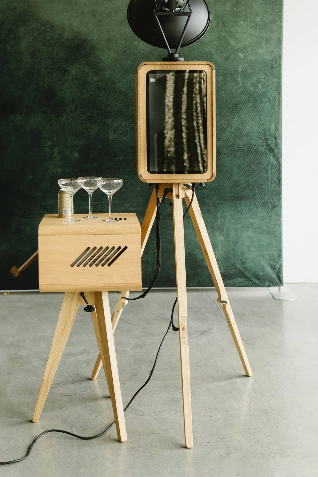 Wooden photo booth on a tripod in front of an emerald green velvet backdrop with champagne coupes on a side table