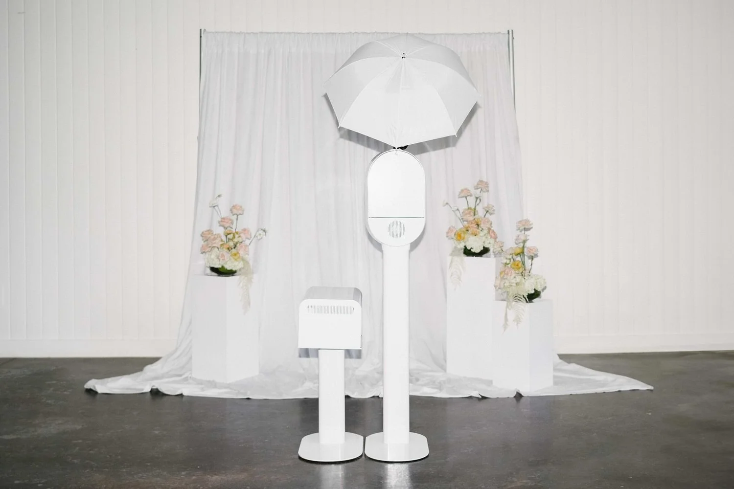 White open-air photo booth with a studio umbrella light in front of a draped white backdrop and florals