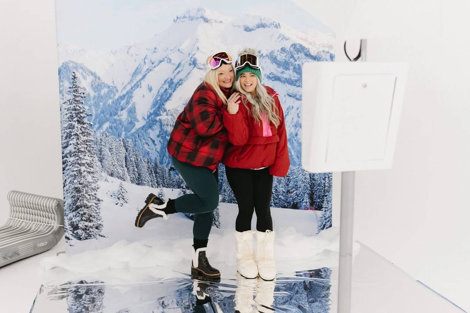 Two guests in plaid and red jackets posing at an open-air photo booth with a snowy mountain backdrop