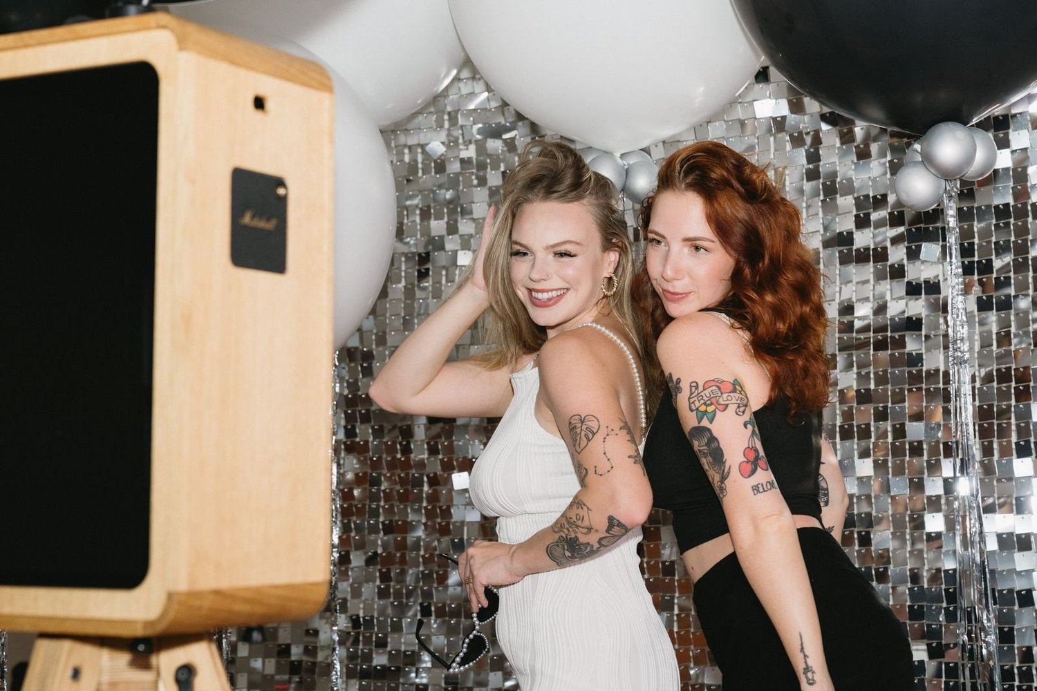 Two friends posing beside a wooden-frame photo booth against a silver sequin wall