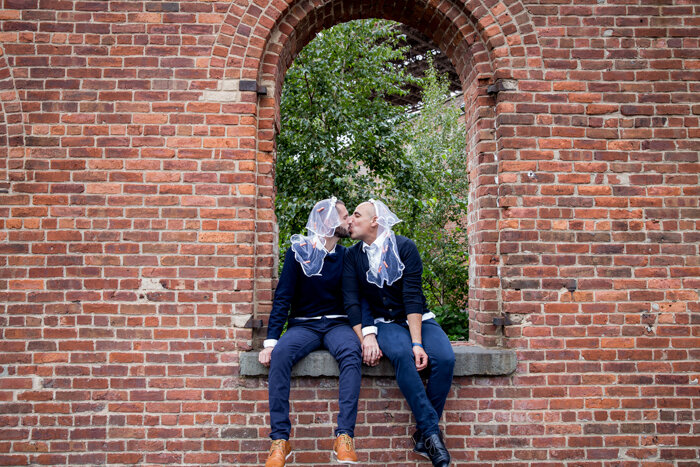 two grooms wearing veils kiss in brick archway after wedding