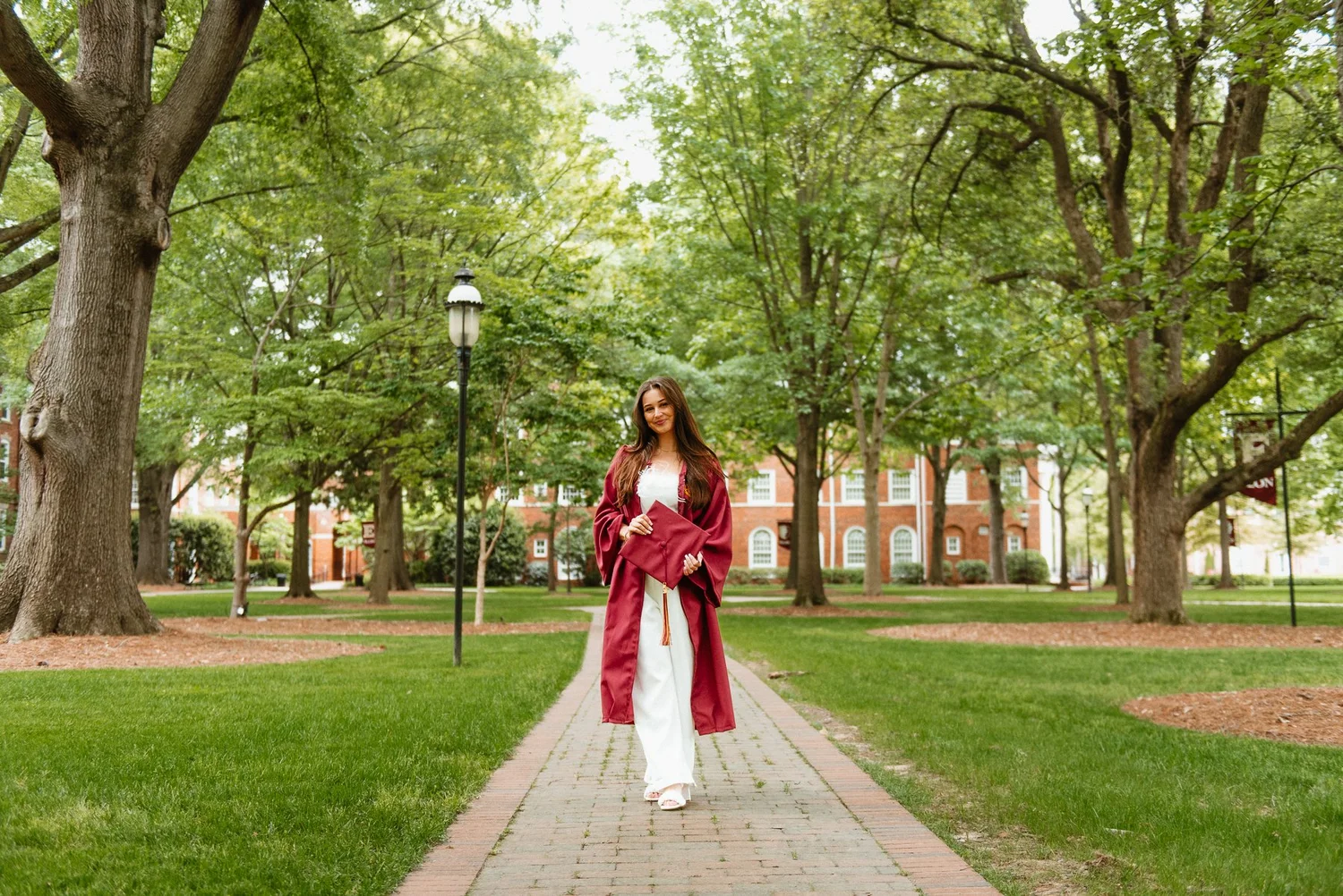 Elon senior in a maroon gown walking a brick path lined with tall oak trees