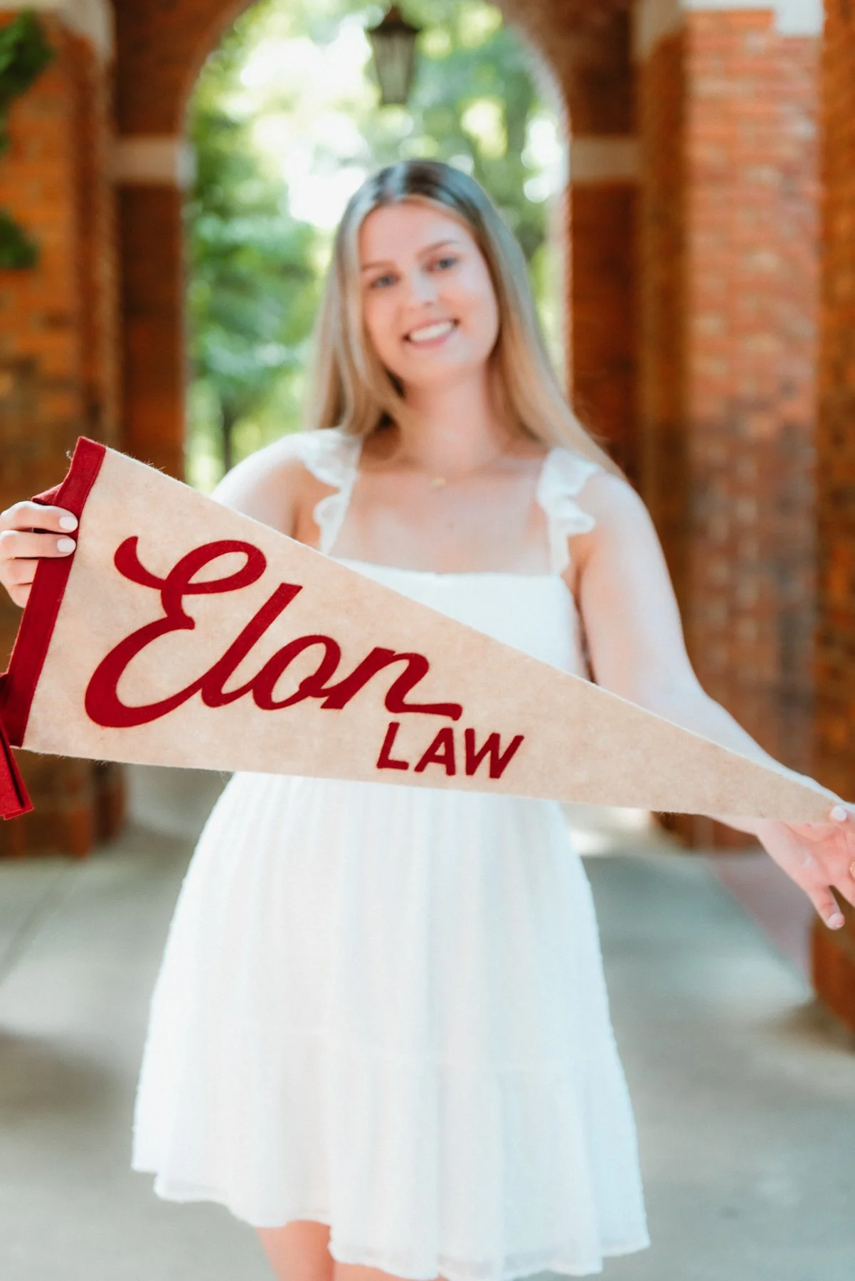 Elon senior in a white dress holding an Elon Law pennant
