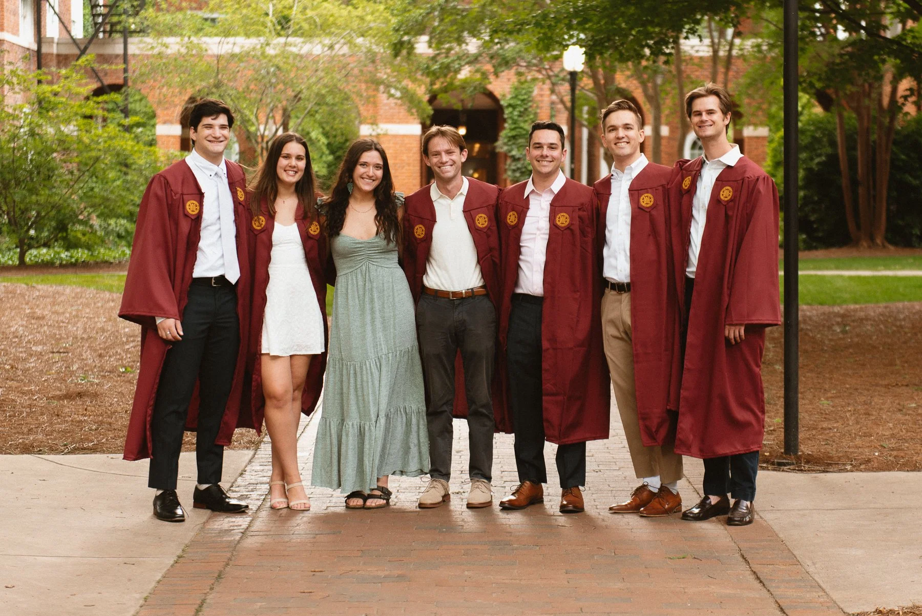 Group of Elon seniors in maroon gowns and friends standing together on a brick path on campus