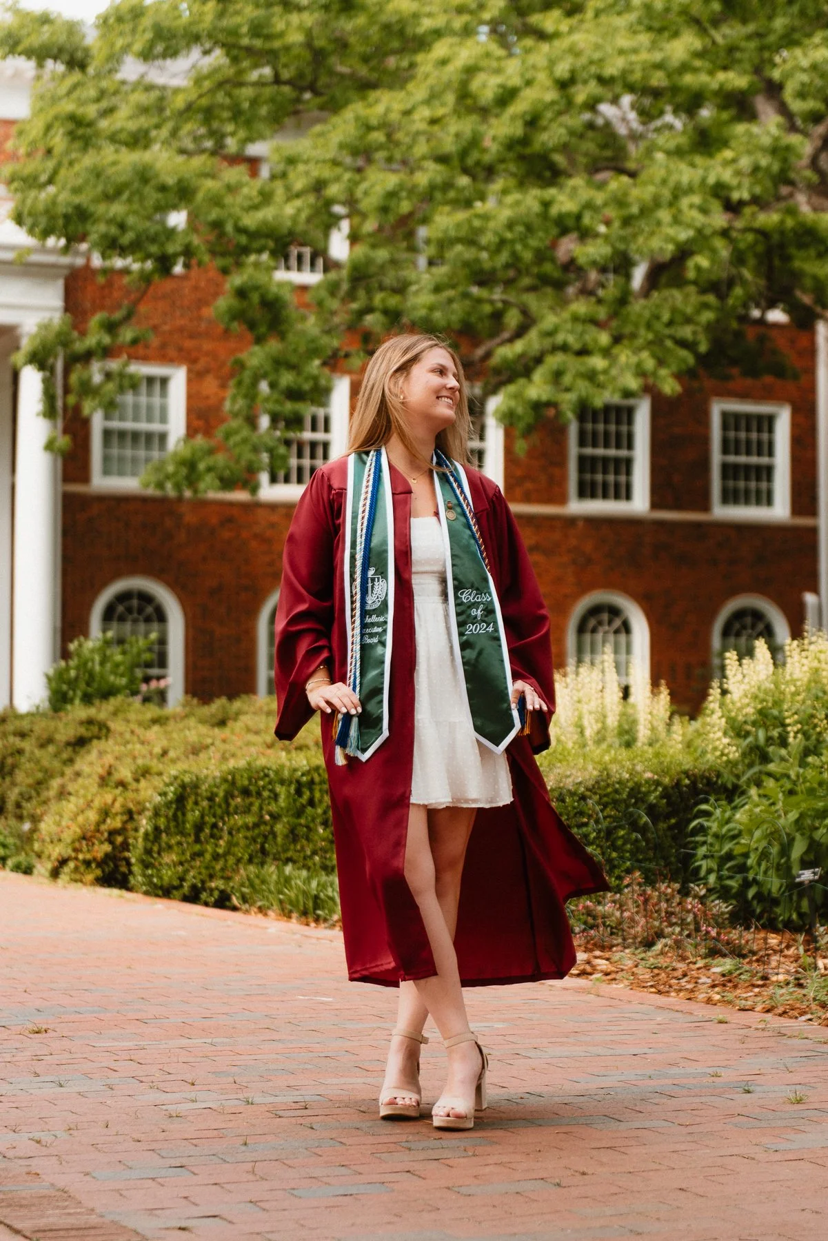 Elon graduate in a maroon gown and stole on a brick walk in front of a residence hall