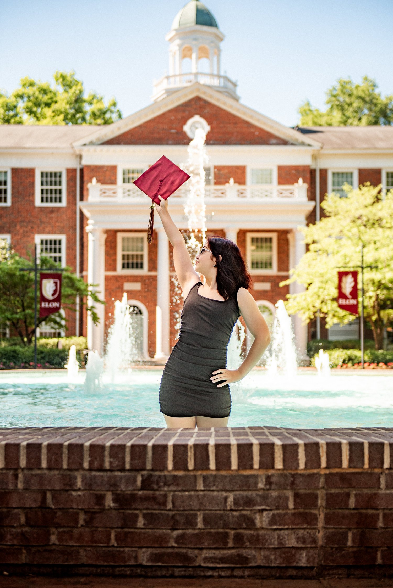 Elon senior holding her cap in the air at the fountain in front of a cupola building