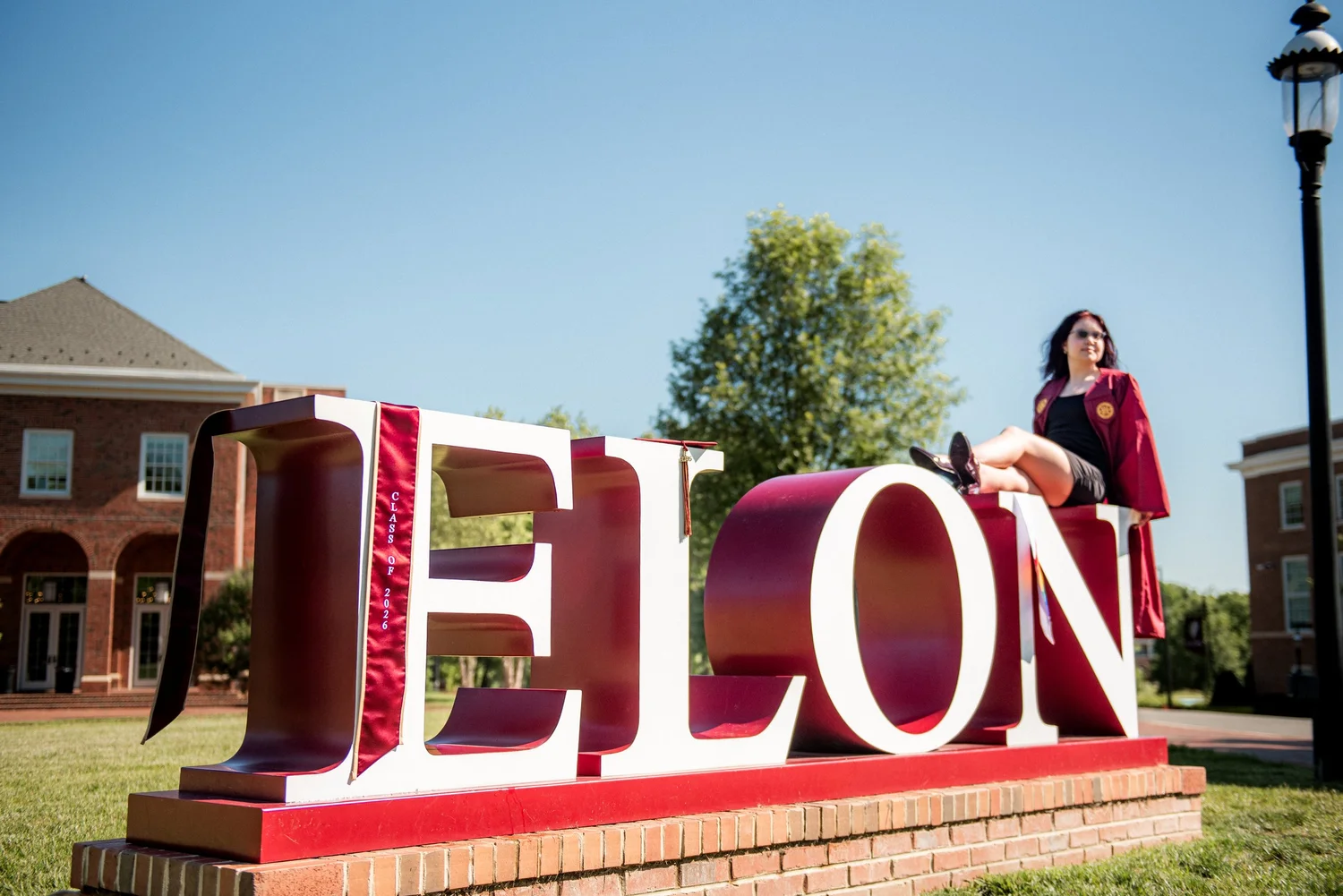 Elon senior in a maroon gown sitting on the large ELON letters on campus