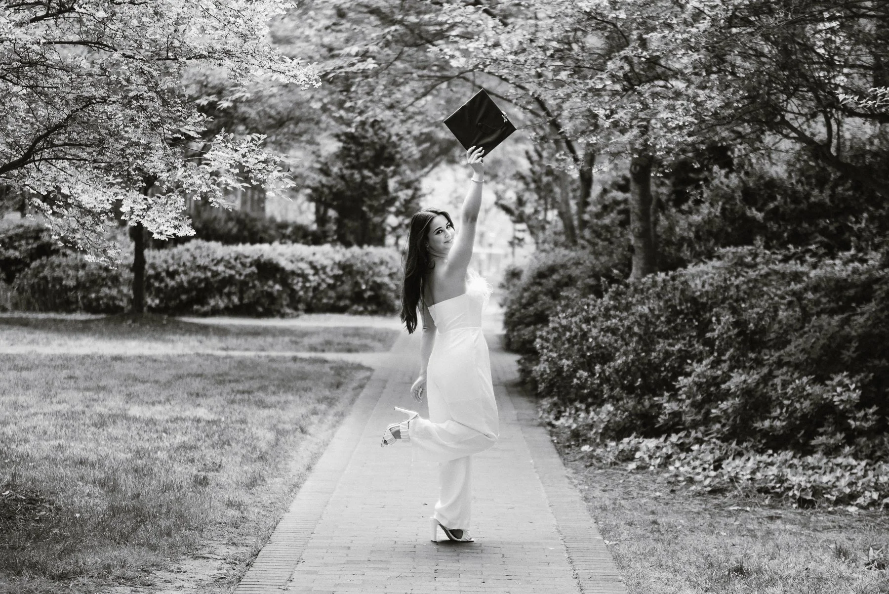 Black and white photograph of an Elon graduate tossing her cap on a tree-lined path