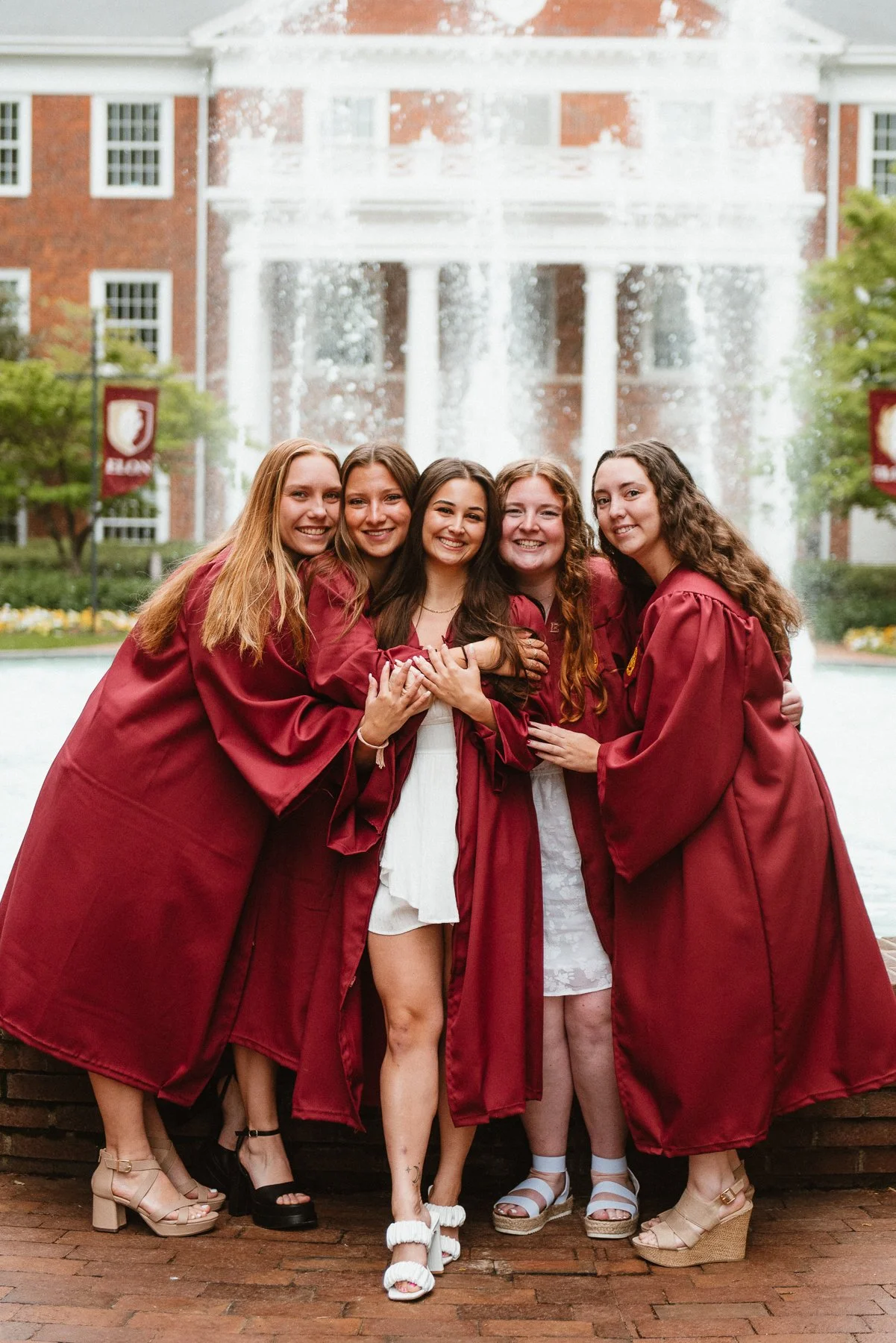 Five Elon seniors in maroon gowns laughing together in front of a fountain