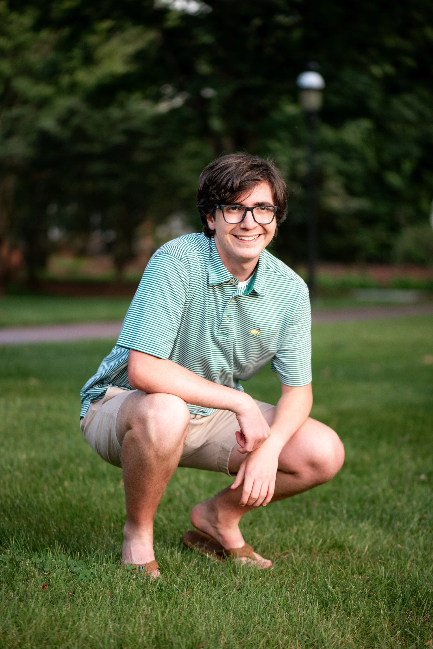 Elon senior in a green polo crouching on the lawn on campus