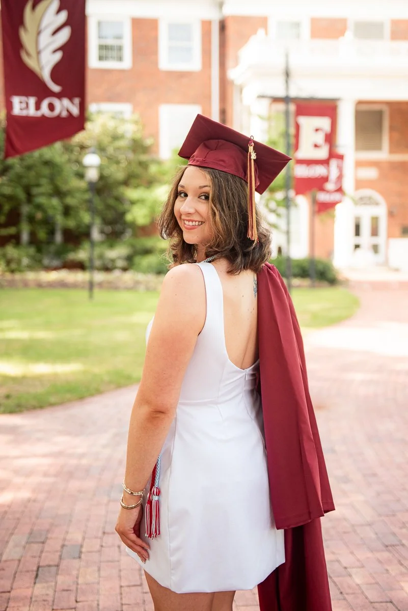 Elon senior in a white dress and maroon gown looking back over her shoulder on campus