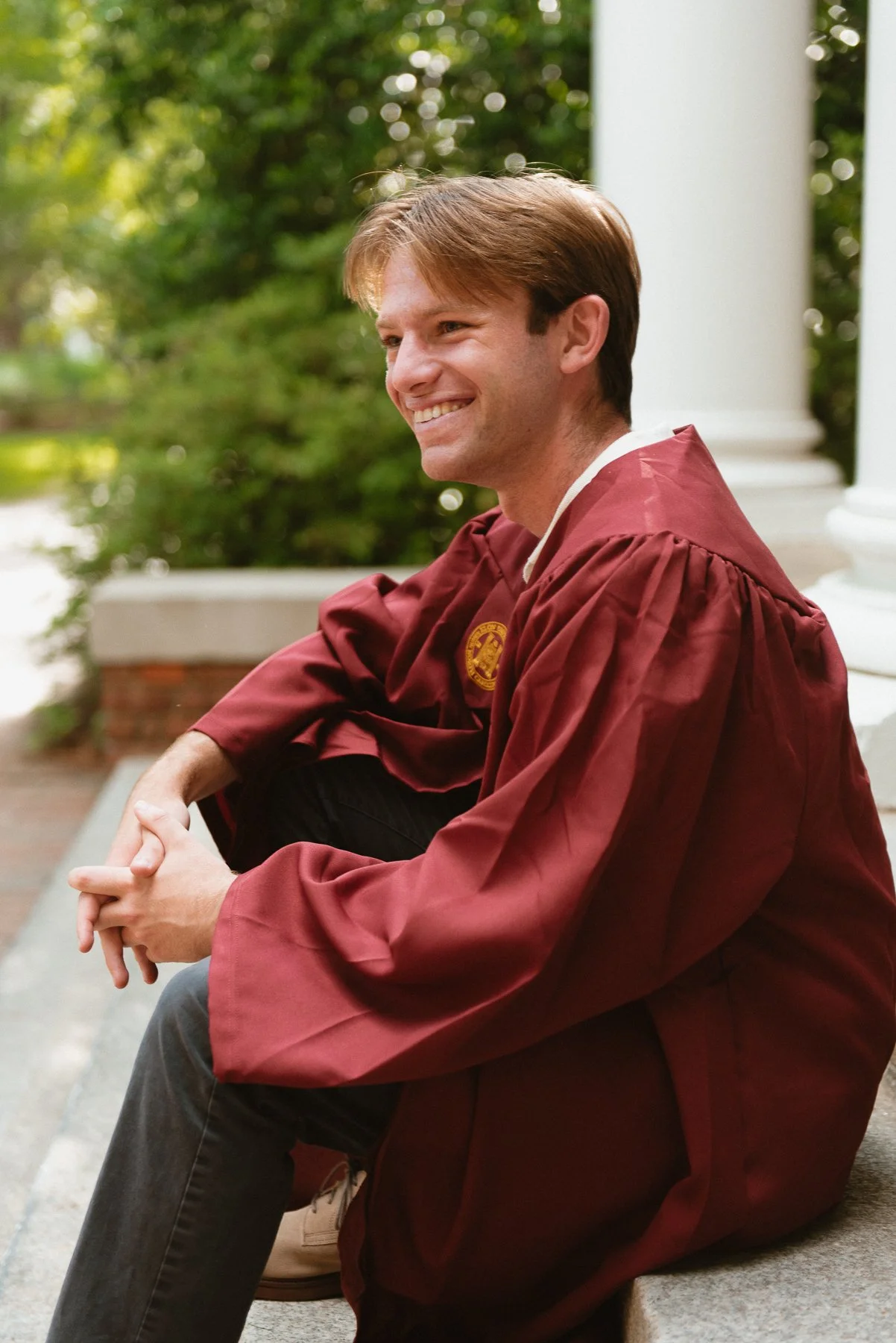 Elon graduate in a maroon gown laughing on the steps beside white columns