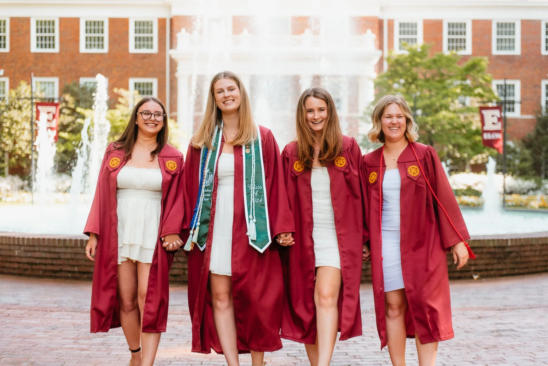 Four Elon seniors in maroon gowns and stoles laughing together at Fonville Fountain in front of the Alamance Building