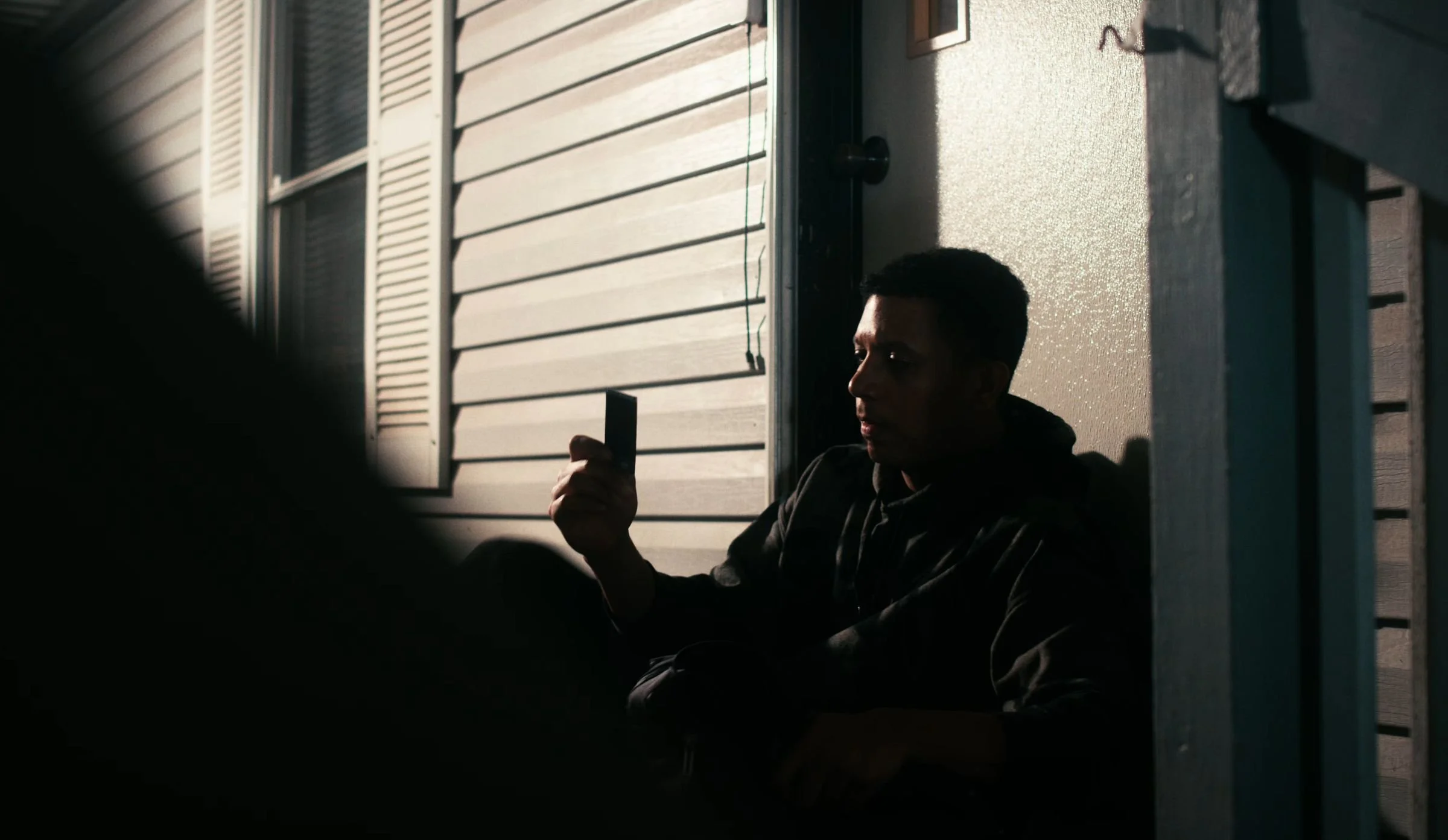 Still from INERTIA: a man in a dark hooded sweatshirt sits against the siding of a house holding up a small dark rectangular object in a beam of light, deep shadow filling the left side.