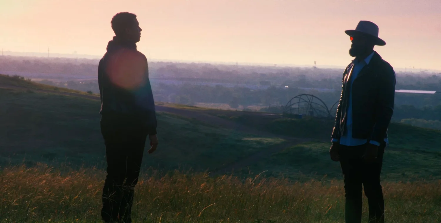 Still from INERTIA: two people stand facing each other in tall grass on a hilltop, one in a hooded sweatshirt and one in a brimmed hat and jacket, a hazy valley behind them.