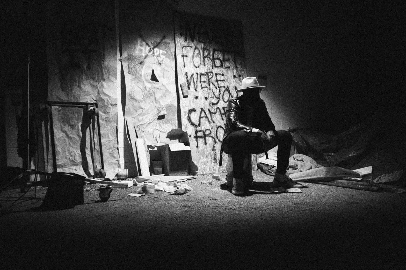 Behind the scenes on INERTIA: an actor sits in a single pool of hard light beside a graffiti wall, the rest of the frame in darkness.