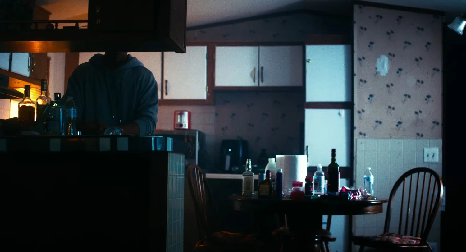 Still from INERTIA: a dim kitchen where a figure in a hoodie stands at a counter lined with liquor bottles, a round table cluttered with bottles.