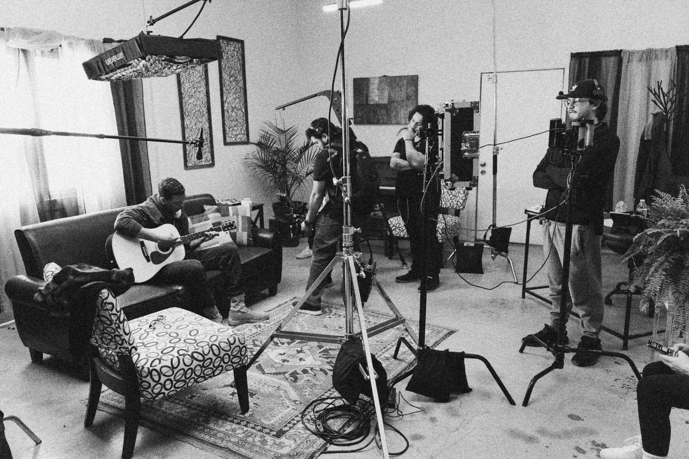 Behind the scenes on INERTIA: wide view of the living room set: actor with guitar on the couch, crew, lights and boom around him.