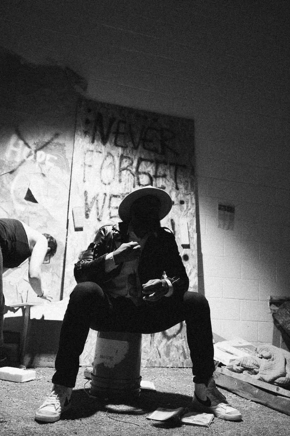 Behind the scenes on INERTIA: an actor in a hat seated on a bucket before a graffiti covered board under hard light.