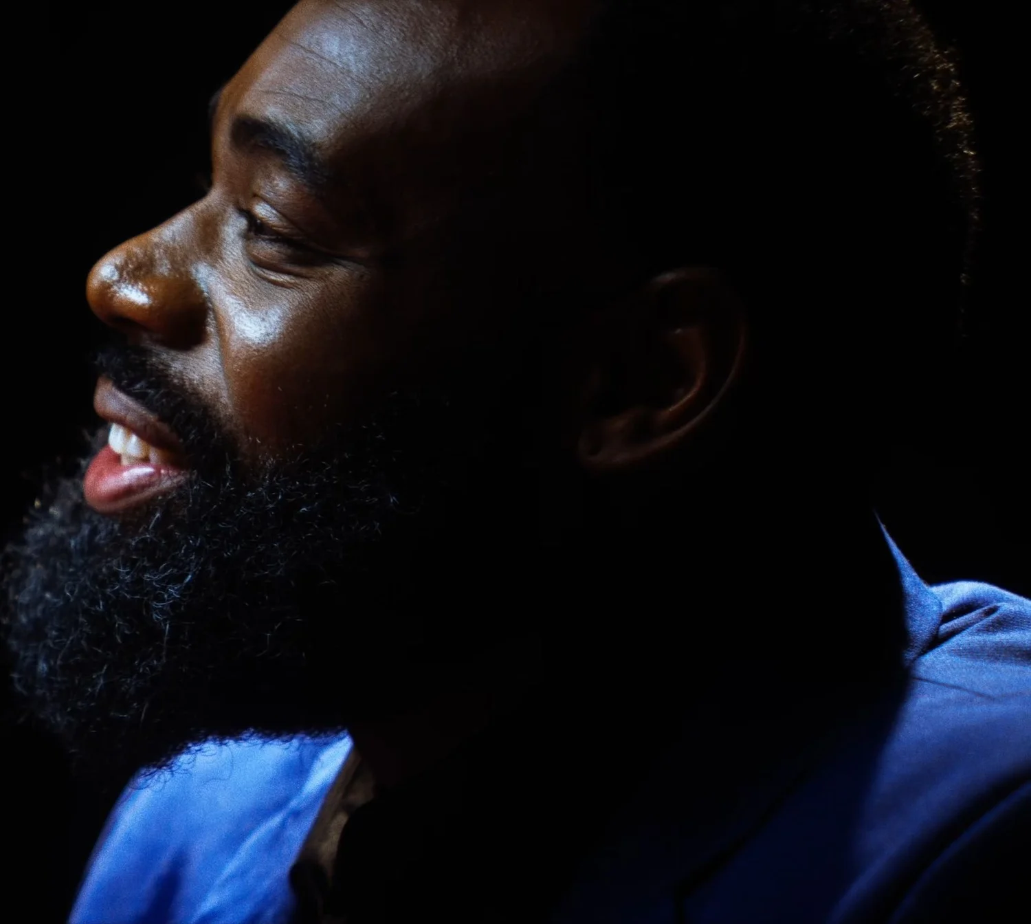 Still from INERTIA: a bearded man in a blue jacket is seen in profile with an open smile, lit from the side against a black background.