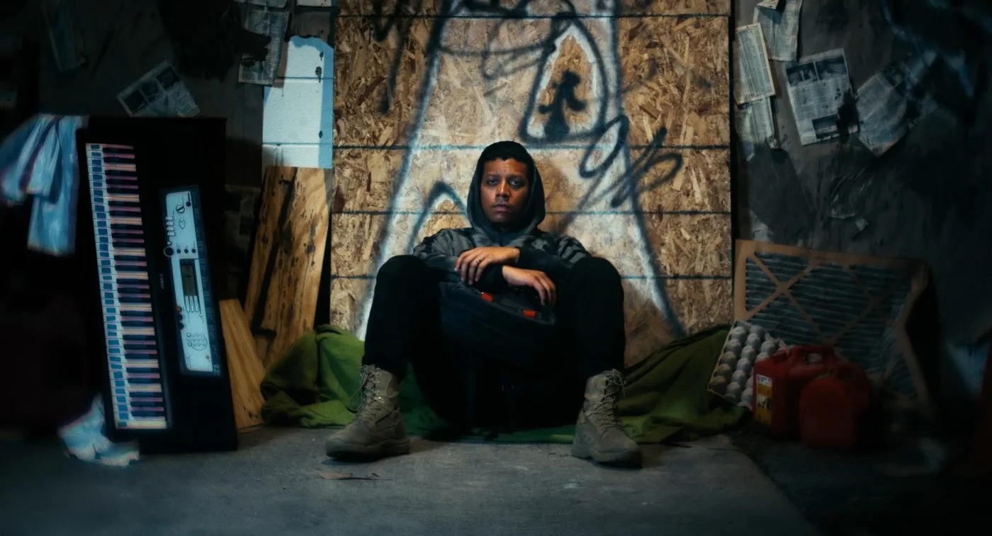 Still from INERTIA: a person in a hooded jacket and boots sits on a blanket against a plywood board covered in graffiti, a dark bag on their lap, with a keyboard to one side and red gas cans to the other.