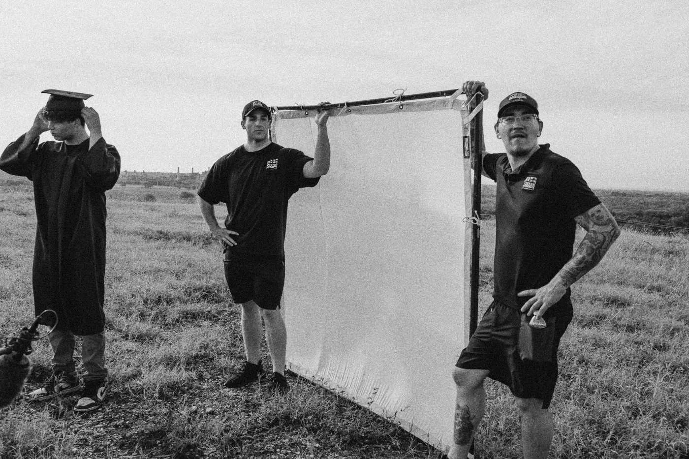 Behind the scenes on INERTIA: crew members hold a large diffusion frame in a field beside a person in a graduation cap and gown.