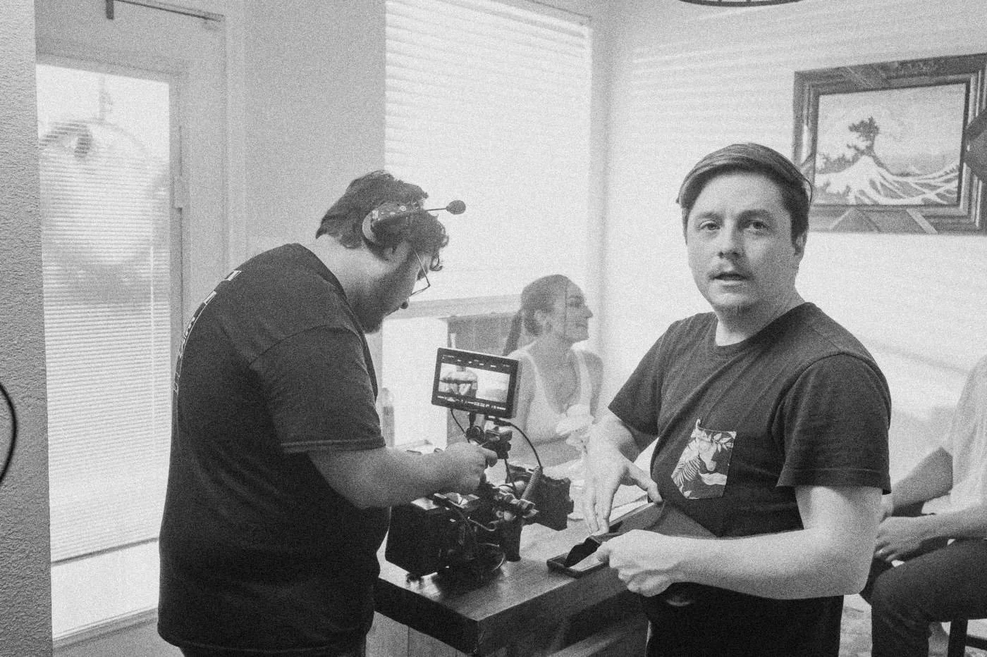 Behind the scenes on INERTIA: a camera operator and a crew member talk beside a table where an actress sits.