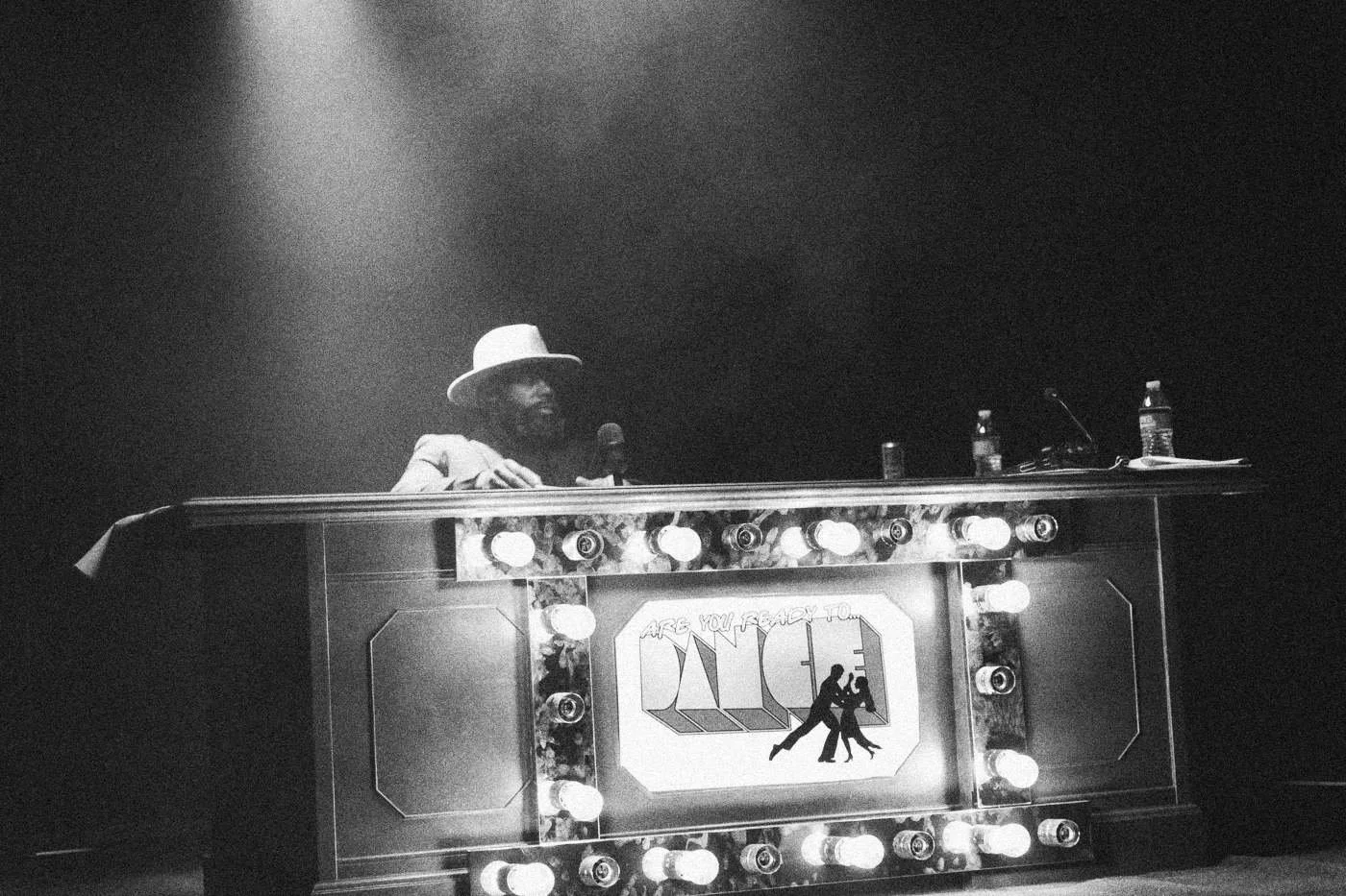 Behind the scenes on INERTIA: an actor in a hat sits behind a lit game show style desk in a hazy spotlight.