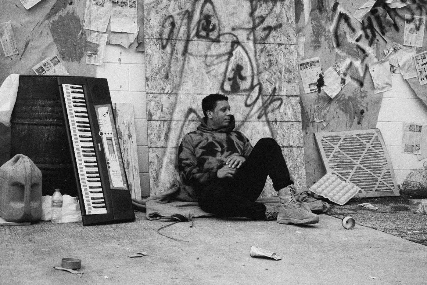 Behind the scenes on INERTIA: an actor in a hoodie sits against a graffiti covered set wall beside a keyboard and pallets.