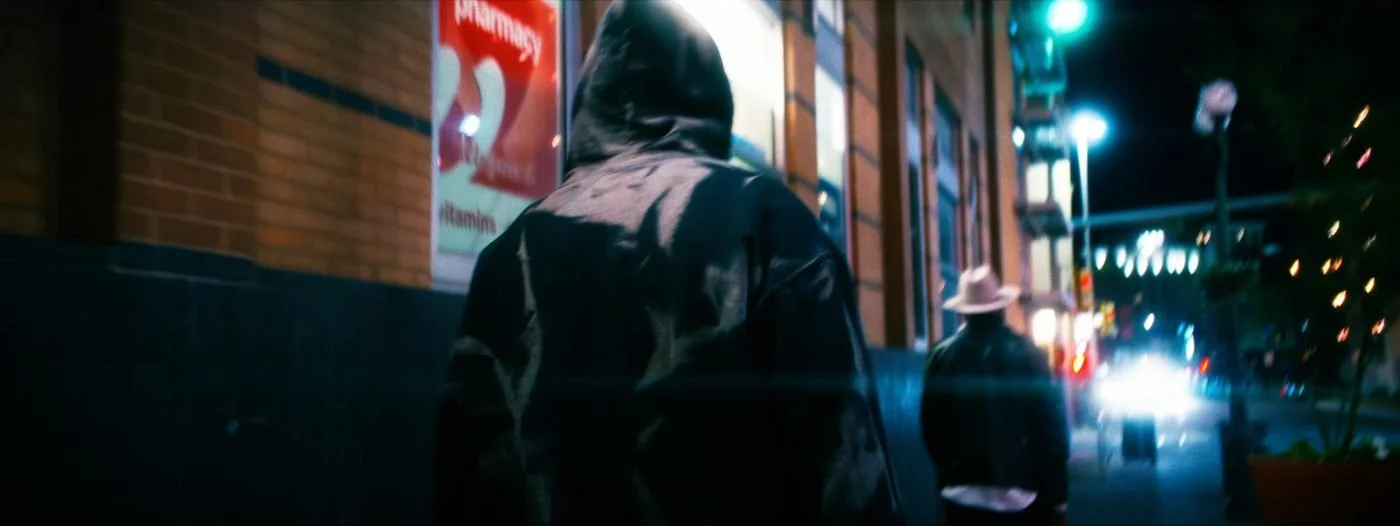 Still from INERTIA: a hooded figure seen from behind walks along a city sidewalk at night past a lit pharmacy sign, a man in a light hat walking ahead.