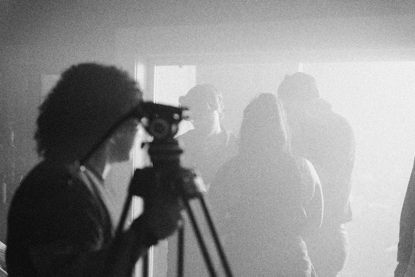 Behind the scenes on INERTIA: a camera operator at a tripod lines up a shot of party cast in pale haze.