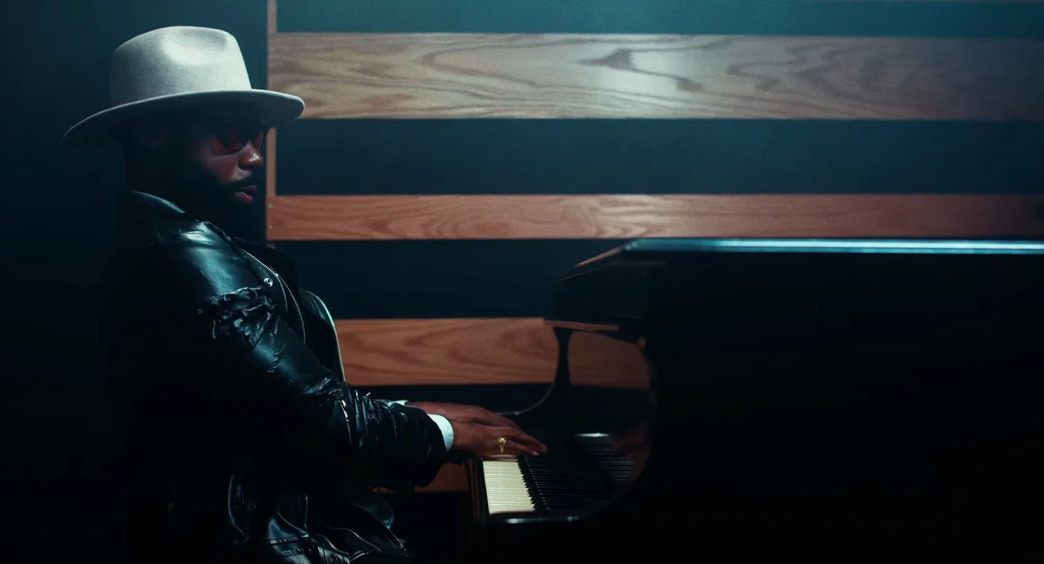 Still from INERTIA: a man in a white wide brimmed hat, tinted glasses and a black leather jacket plays a grand piano in front of a wall of horizontal wood slats.