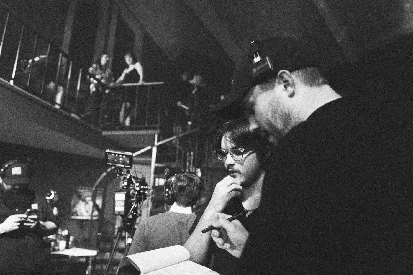 Behind the scenes on INERTIA: a crew member in a cap reads notes with a colleague while a camera team works on the balcony stairs behind.