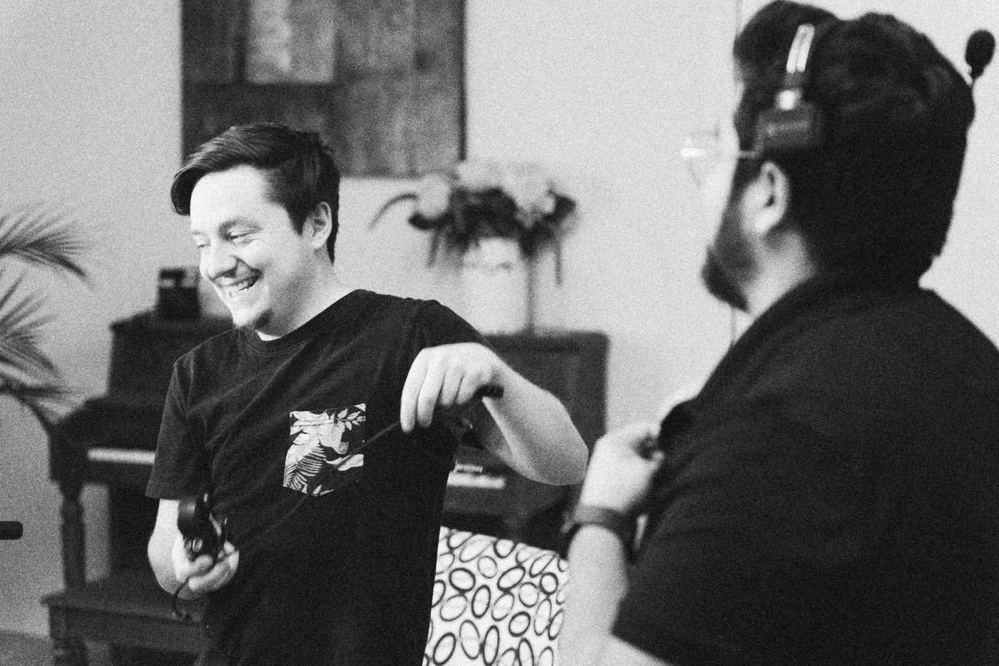 Behind the scenes on INERTIA: a crew member laughs while holding a microphone, talking with a colleague on the living room set.