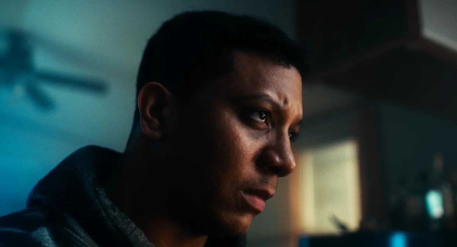 Still from INERTIA: a close view of a man in a dark hooded sweatshirt in profile, the shadow of a ceiling fan on the wall behind him.