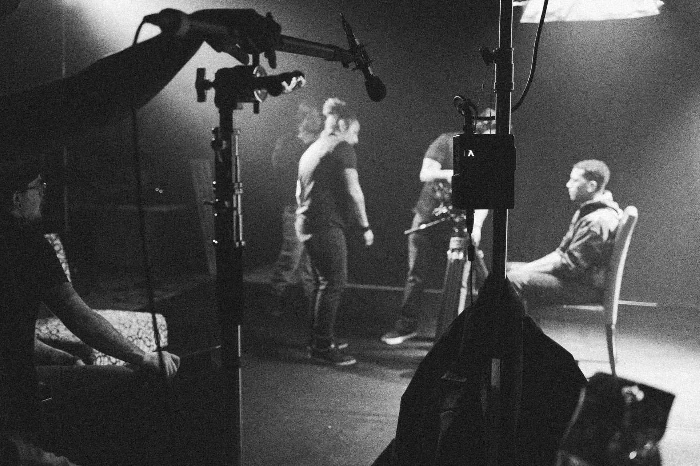 Behind the scenes on INERTIA: wide view of a dark studio with light stands, a boom and crew around an actor in a red chair.