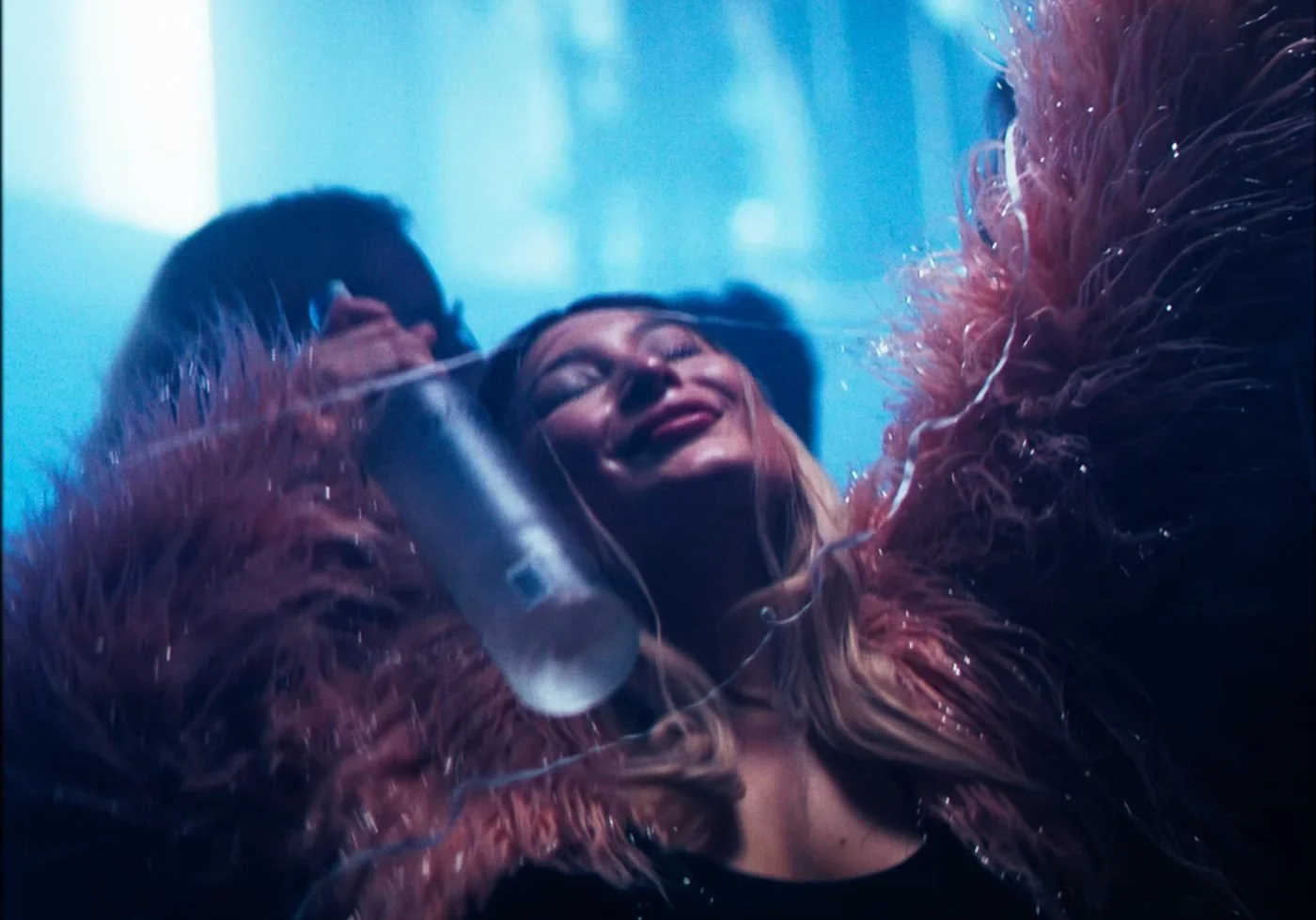 Still from INERTIA: a woman in a pink feathered coat tilts her head back with her eyes closed, holding a bottle beside her face under blue light, a person behind her.
