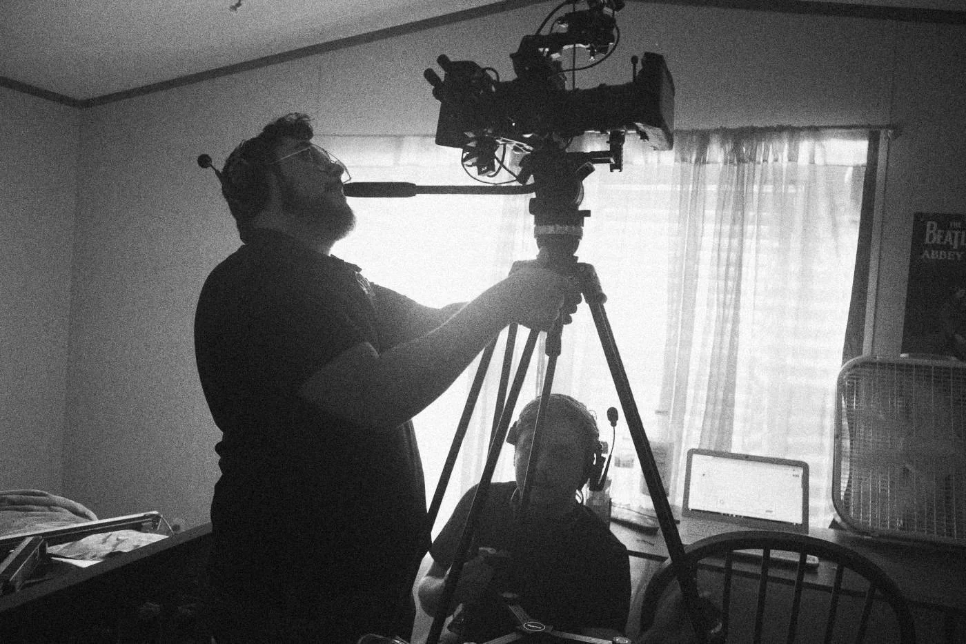 Behind the scenes on INERTIA: a camera operator on a tripod stands in silhouette against a curtained window, a colleague seated below.