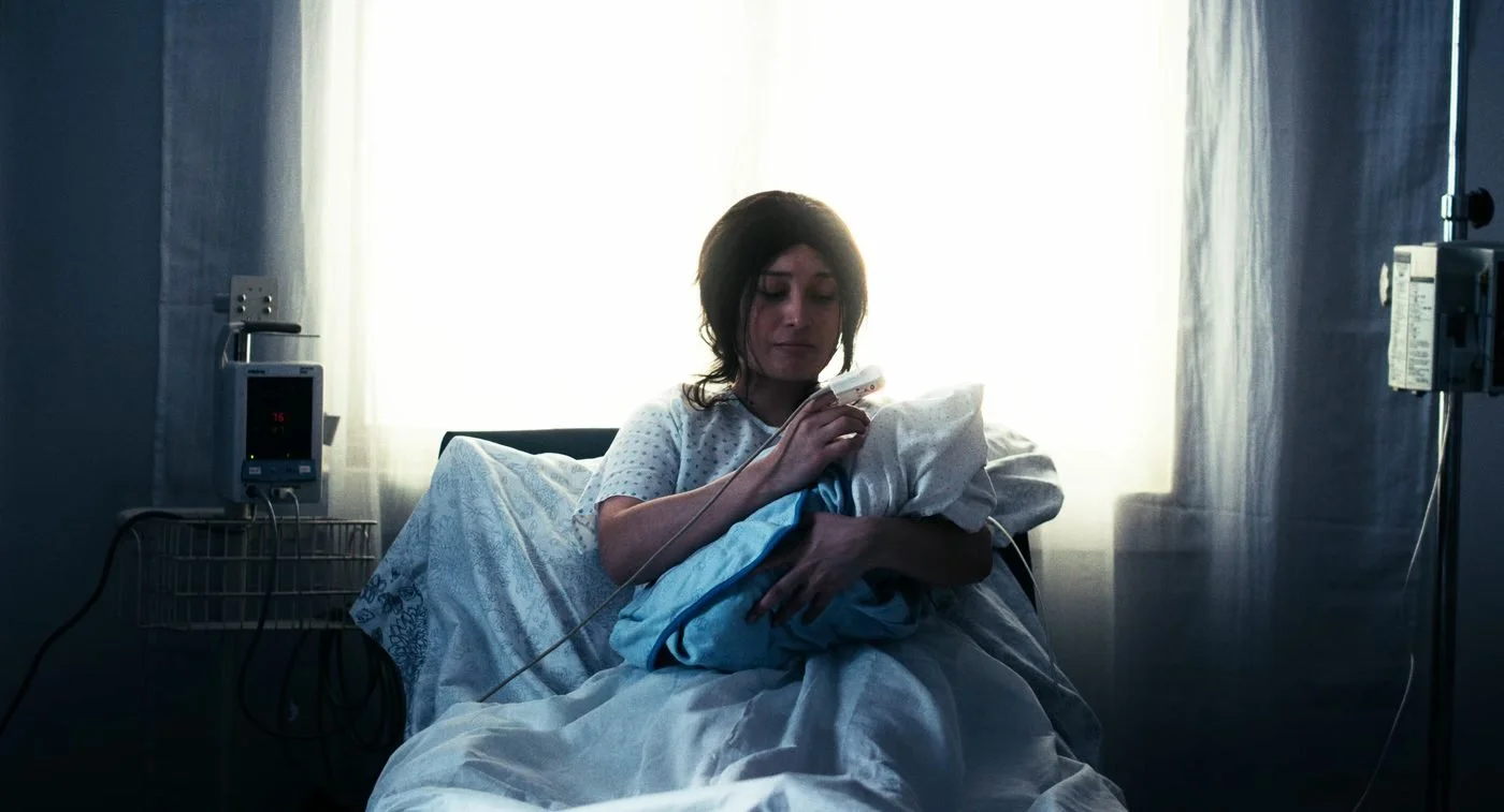 Still from INERTIA: a woman in a hospital gown sits in a hospital bed holding a bundle wrapped in a blue blanket, a monitor clip on her finger and a bright curtained window behind her.