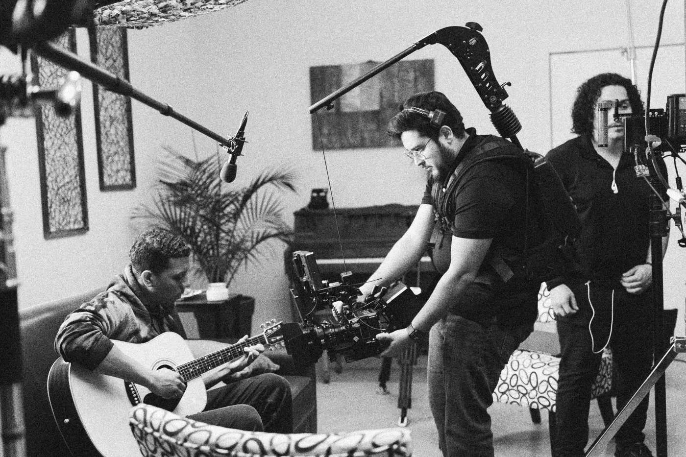 Behind the scenes on INERTIA: a camera operator with a handheld rig films an actor playing guitar on a couch while a boom operator listens.