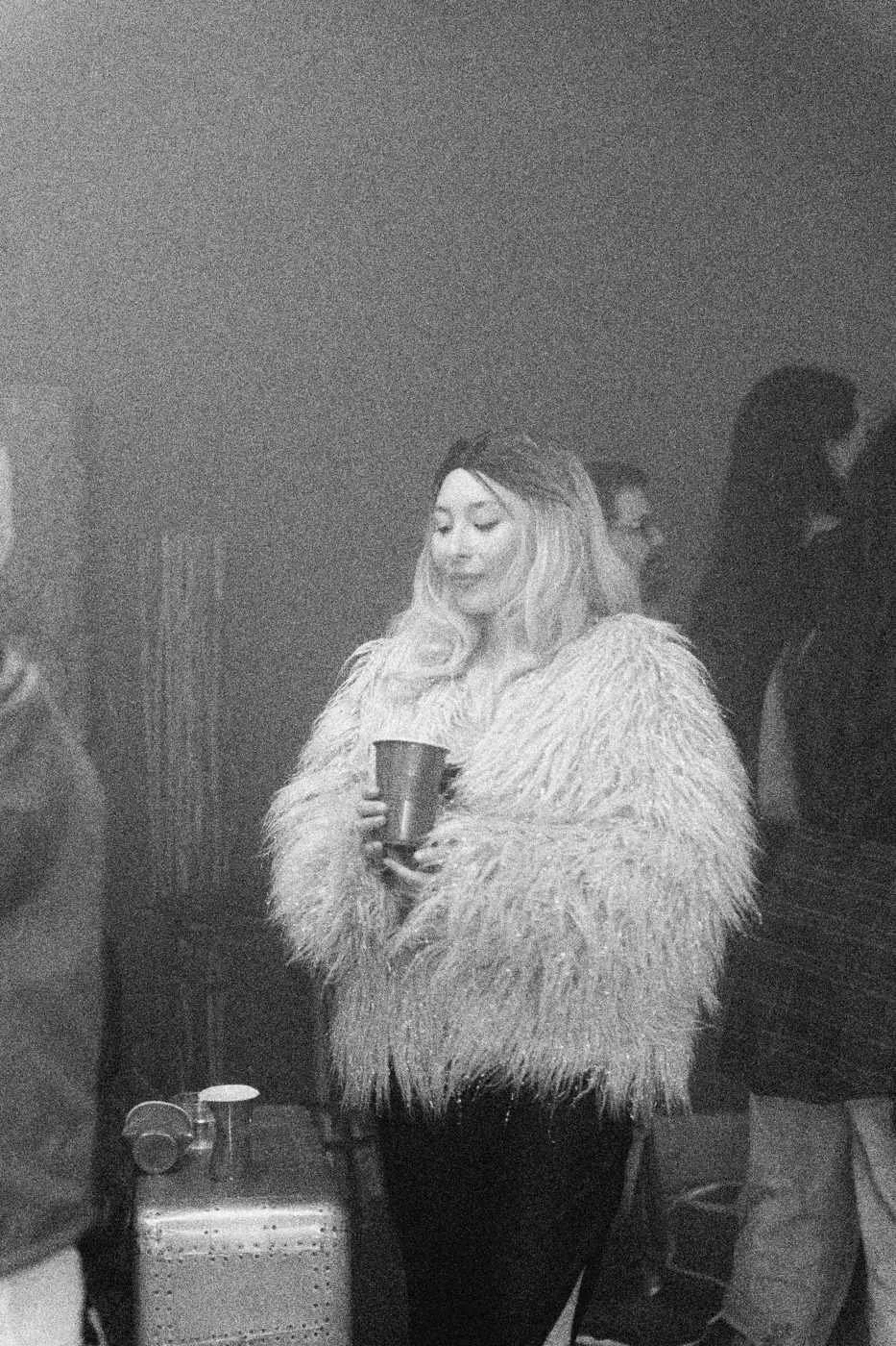 Behind the scenes on INERTIA: an actress in a pink faux fur coat holding a red cup at a hazy party.