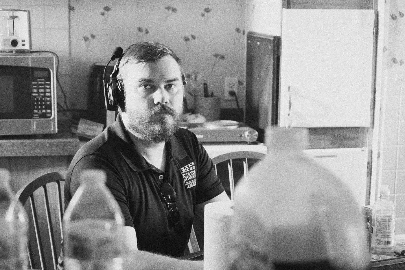 Behind the scenes on INERTIA: a bearded crew member in a headset sits at a kitchen table, water bottles in the foreground.