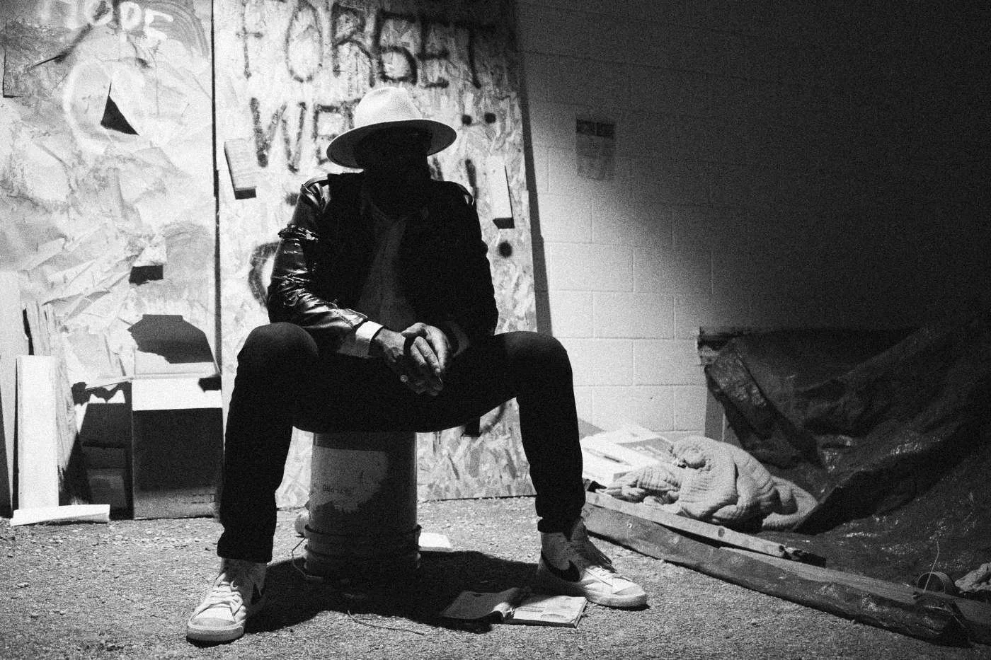 Behind the scenes on INERTIA: an actor in a hat and leather jacket sits on a bucket before a board painted NEVER FORGET, lit by hard light.