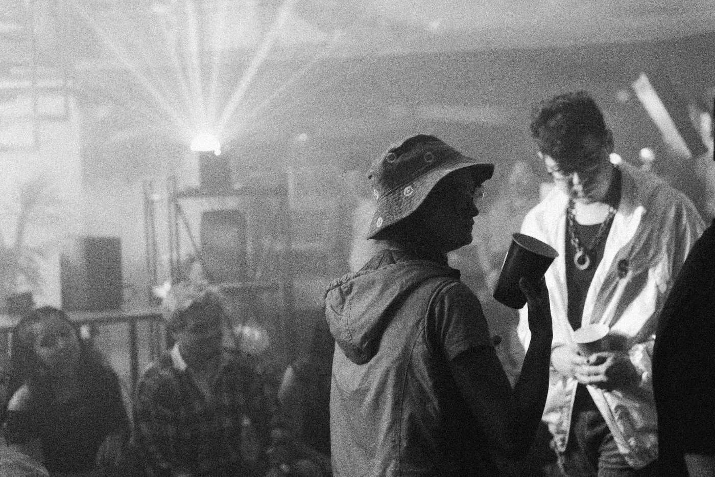 Behind the scenes on INERTIA: party guests in haze and green laser light, one in an orange bucket hat holding a red cup.