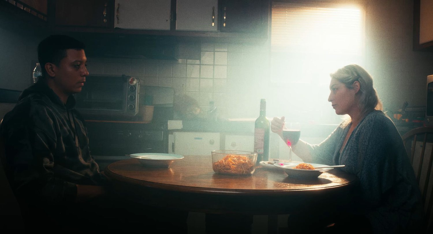 Still from INERTIA: a man in a dark hooded sweatshirt and a woman holding a glass of red wine sit across a round kitchen table with bowls of spaghetti and a wine bottle, hazy sunlight coming through a window.