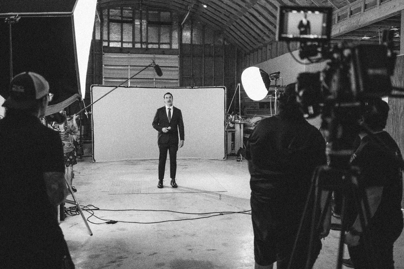 Behind the scenes on INERTIA: wide view of a warehouse studio where an actor in a suit stands before a green screen, camera and crew in the foreground.